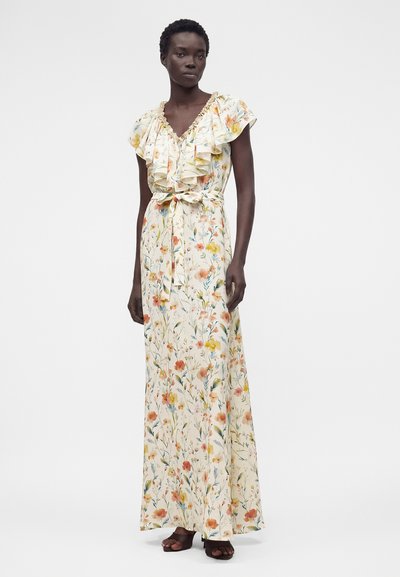 Γυναίκα με αμάνικο, κυματιστό, φλοράλ μάξι φόρεμα με δεμένη ζώνη στη μέση και μαύρα ανοιχτά παπούτσια, στέκεται μπροστά σε απλό φόντο.