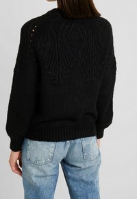 Zwarte gebreide trui met een gestructureerd patroon op de achterkant, geribbelde boorden en zoom. Gepaarde met lichtblauwe denim jeans.