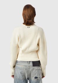 Suéter de canalé crema con cuello redondo y hombros caídos, que presenta un pequeño acento negro en la parte posterior. Combinado con jeans desgastados y descoloridos.