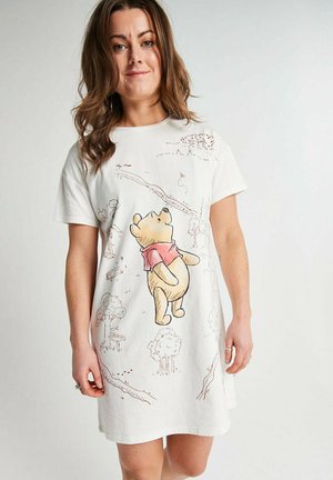 Woman wearing white short-sleeve dress featuring Winnie the Pooh illustration walking among trees and paths on the fabric.
