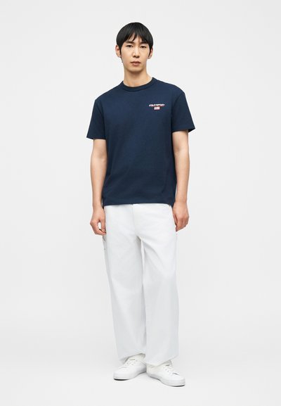 Νέος άνδρας που στέκεται φορώντας μπλε σκουρόχρωμο μπλουζάκι Polo Sport, άνετο λευκό παντελόνι και λευκά αθλητικά παπούτσια, μπροστά σε απλό φόντο.