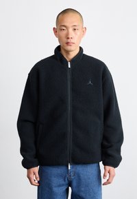 Giacca in pile nera con zip frontale, colletto alto e tasche laterali. Presenta un piccolo logo sul petto. Tessuto morbido e strutturato.