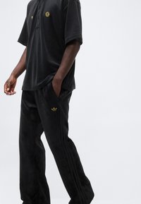 Mann trägt schwarze Adidas-Trackpants aus Velours und ein passendes schwarzes Polohemd mit kurzen Ärmeln und goldenen Logos, steht vor einem einfarbigen Hintergrund.