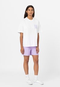 Vit bomull t-shirt med lila text på vänster bröst, matchad med ljuslila shorts. Modellen bär vita sneakers och ankelsockor.