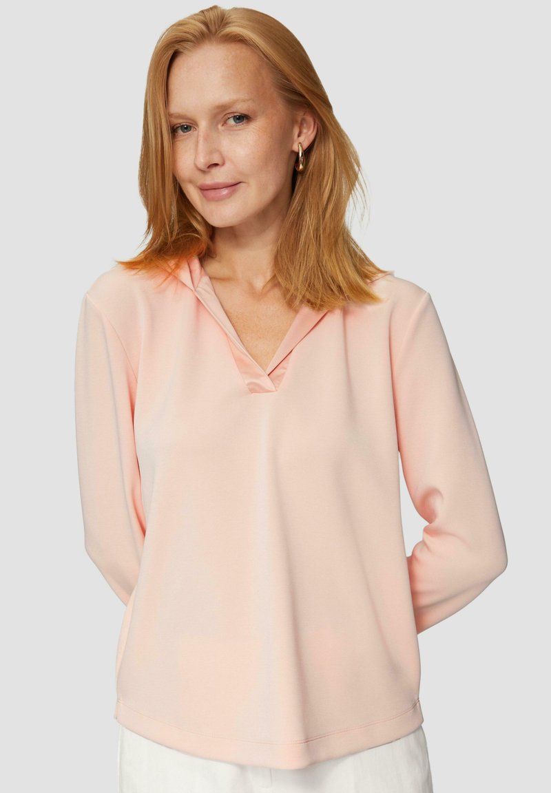 Vrouw met steil rood haar, gekleed in gouden ringoorbellen en een lichtroze blouse met lange mouwen, die lichtjes glimlacht tegen een grijze achtergrond.