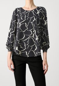 Chemisier noir avec des motifs abstraits blancs irréguliers, manches longues froncées, porté avec un pantalon noir, section médiane échancrée sur fond uni.