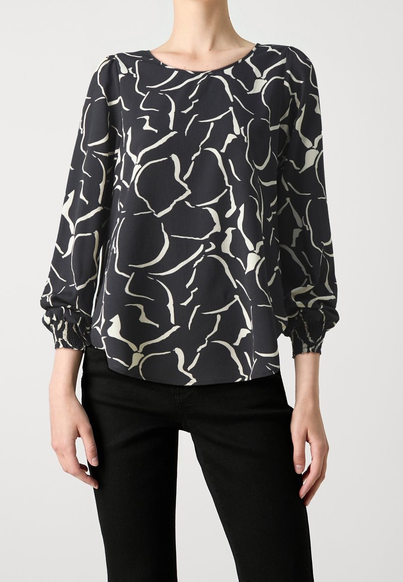 Chemisier noir avec des motifs abstraits blancs irréguliers, manches longues froncées, porté avec un pantalon noir, section médiane échancrée sur fond uni.