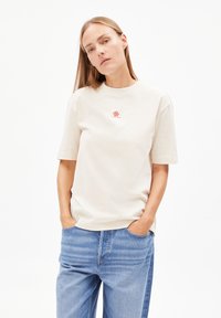 Beige katoenen t-shirt met korte mouwen, ronde halslijn en een rood geborduurd bloemenlogo op de borst. Gepaard met lichtblauwe spijkerbroek.