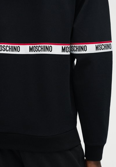 Μαύρο πουκάμισο μακρυμάνικο με λευκές οριζόντιες λωρίδες στο στήθος και το χέρι, που φέρουν επαναλαμβανόμενο μαύρο κείμενο "MOSCHINO" και λεπτή κόκκινη ρίγα πάνω από αυτές.