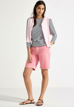 Ung kvinde iført lyserøde knælange shorts, sort-hvidstribet langærmet top, lyserød sweater draperet over skuldrene og brune sandaler.