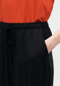 Άτομο που φοράει μαύρο παντελόνι με κορδόνι στη μέση, με το χέρι στην τσέπη και πορτοκαλί μπλουζάκι φορεμένο μέσα στη μέση, μπροστά σε απλό φόντο.