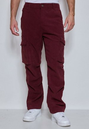 Homme portant un pantalon cargo en velours côtelé bordeaux et des baskets blanches, debout devant un fond clair uni.