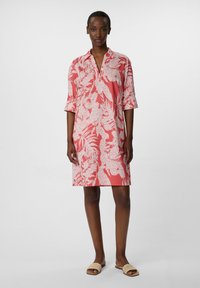 Koronkowa sukienka w kolorze koralowym z kołnierzykiem, rękawami o długości trzech czwartych i białym kwiatowym wzorem. Gładki materiał i długość do kolan. Noszona z beżowymi klapkami.