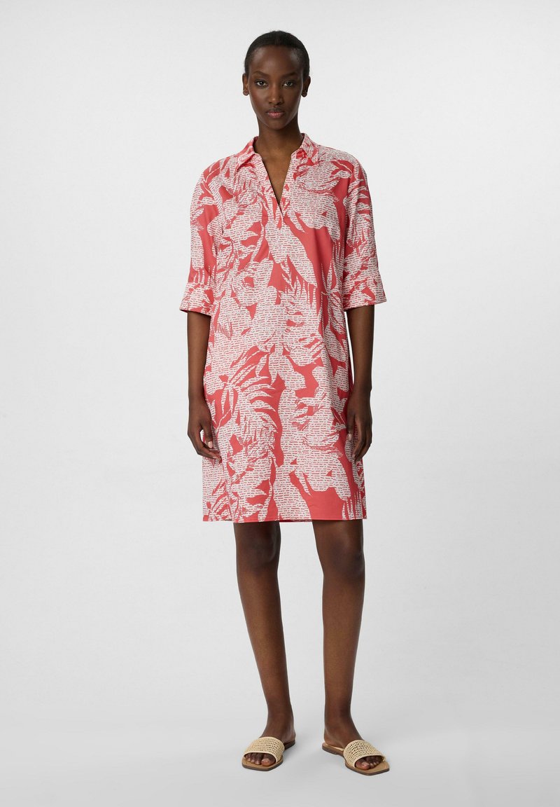 Koronkowa sukienka w kolorze koralowym z kołnierzykiem, rękawami o długości trzech czwartych i białym kwiatowym wzorem. Gładki materiał i długość do kolan. Noszona z beżowymi klapkami.