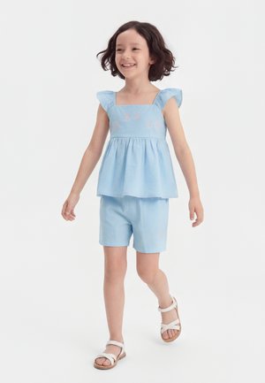 Mladé dievča sa usmieva, kráča, má na sebe svetlomodrý tielko bez rukávov s volánmi, zodpovedajúce šortky a biele sandále na bielom pozadí.