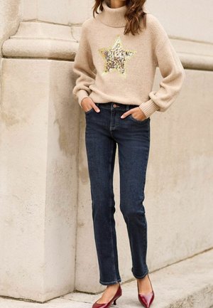 Pull en maille beige avec un motif d'étoile métallique, associé à un jean bleu foncé et des escarpins rouges. Tissu texturé et coupe décontractée.