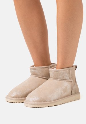 Bottes beige scintillantes à la cheville avec une texture douce, un bout rond et des accents surpiqués. Dotées d'un col rabattu et d'une semelle en caoutchouc plate.