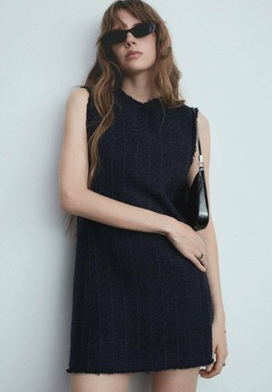 Navyblaues ärmelloses Kleid aus texturiertem Stoff, mit einem fransigen Saum und vertikalen Streifen. Kombiniert mit einer kleinen schwarzen Handtasche.