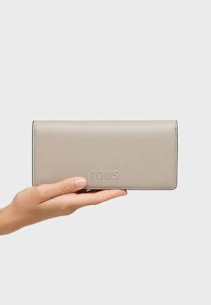Mano sosteniendo una cartera de cuero beige con el relieve "TOUS" sobre un fondo liso de color gris claro.