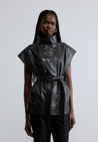 Zwart lederen vest met hoge kraag, korte mouwen en een taille met ceintuur. Voorzien van geribbelde accenten en knopen aan de voorkant. Soepele textuur.