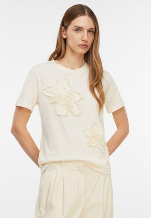 Donna che indossa una maglietta crema a maniche corte con due applicazioni floreali testurizzate e pantaloni plissettati di colore chiaro abbinati.