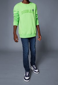 Neonowa zielona bluza z nadrukiem "DOUBLE", niebieskie dżinsy oraz granatowo-białe trampki. Bluza ma okrągły dekolt i długie rękawy.