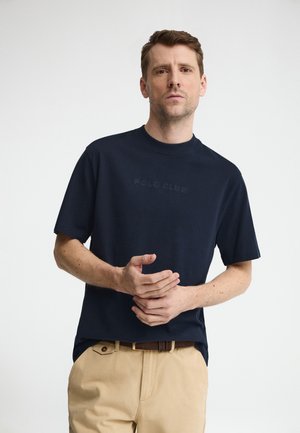 Uomo dalla pelle chiara e capelli castani corti, che indossa una maglietta blu navy "Polo Club" e pantaloni beige, in piedi davanti a uno sfondo bianco.