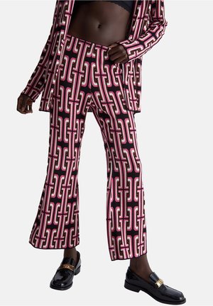 Modèle portant un pantalon à motif géométrique noir, rose et blanc et une veste assortie avec des mocassins noirs sur fond blanc.
