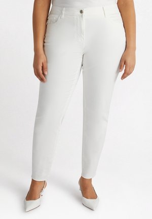 Kvinde iført hvide skinny jeans med høj talje og hvide spidse slingback-hæle på hvid baggrund.