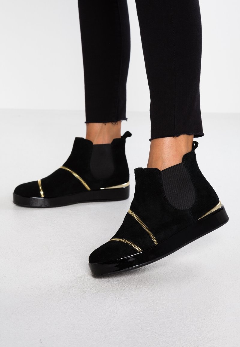 Bottines en daim noir avec détails de fermeture éclair dorée portées avec un pantalon noir, présentées sur un fond de sol blanc.