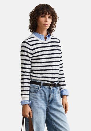 Person med krøllet hår iført en hvid og sort stribet sweater over en blå skjorte, lyseblå jeans og holder en brun taske strop.