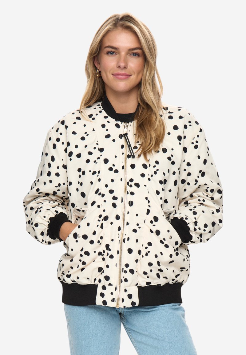 Femme portant une veste blanche zippée avec des pois noirs irréguliers, des poignets et un col noirs, et un jean bleu clair, les mains dans les poches.