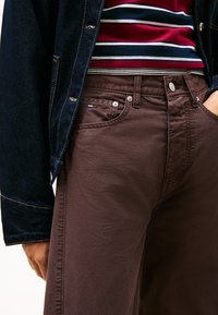 Pantalones de algodón marrón con textura suave y ajuste slim, con un bolsillo delantero y un pequeño detalle de logo. Combinados con una camisa a rayas.