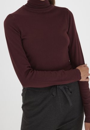 Kobieta ubrana w bordowy sweter z długim rękawem i golfem oraz ciemnoszare spodnie z wiązanym paskiem, stojąca na jednolitym tle.