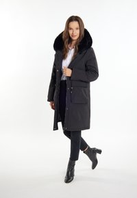 Parka nera con colletto in pelliccia sintetica, chiusura con zip e bottoni, tasche frontali e design a lunghezza media, abbinata a pantaloni neri e stivali alla caviglia.