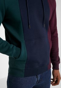 Sudadera a bloque de color que presenta secciones en verde, azul marino y burdeos. Fabricada en un tejido suave, con un bolsillo frontal y capucha con cordones. Ajuste clásico.