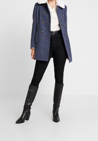 Kvinna i en blå texturerad dubbelknäppt kappa med vit pälskrage, vit blus, svarta högmidjade byxor, svart bälte och knähöga svarta stövlar.