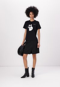Tricou negru din bumbac cu un desen animat cu o pisică purtând ochelari de soare. Asortat cu o fustă neagră, cizme negre până la gleznă și purtând o geantă rotundă neagră.