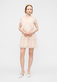 Φόρεμα με βαθμιδωτές στρώσεις από απαλό ροζ τούλι, με βολάν στη λαιμόκοψη και τα μανίκια. Το μοντέλο φοράει λευκές γόβες και στέκεται μπροστά σε απλό φόντο.