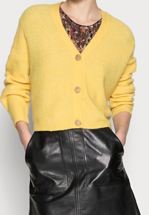 Kvinde iført en gul knap-cardigan over en bluse med blomsterprint, kombineret med et sort lædernederdel, hænderne i lommerne.