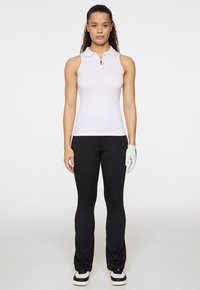 Vit ärmlös pikétröja med dragkedja och krage, kombinerad med svarta åtsittande byxor och en vit handske. Modellen står framåt.