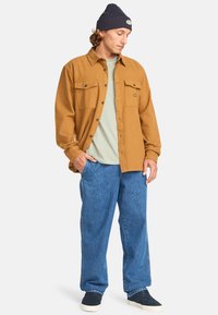 Brun skjorta med knappar och två bröstfickor, lagd över en ljusgrön t-shirt, tillsammans med lösa blå jeans och en marinblå mössa.