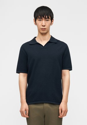 Moški v teksturirani temno modri polo majici s kratkimi rokavi in olivno zelenih hlačah stoji pred enobarvno svetlo ozadje.