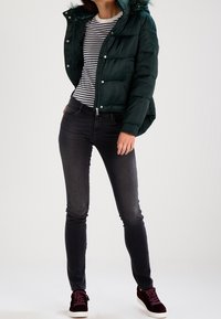 Giacca verde imbottita con cappuccio foderato in pelliccia, maglietta a righe, jeans scuri e sneakers bordeaux. Texture liscia e design aderente.