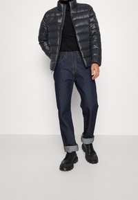 Chaqueta acolchada gris oscuro con cuello alto, llevada sobre un suéter negro de cuello alto, combinada con jeans de mezclilla azul oscuro remangados y botas negras.