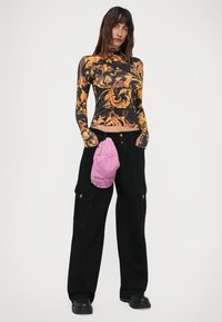 Ženska v črnih cargo hlačah, s črno-oranžno vzorčasto dolgimi rokavi, drži roza kapo "Versace Jeans Couture" pri pasu.