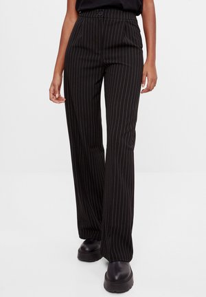 Femme portant un pantalon noir à fines rayures, taille haute et jambes larges, associé à des chaussures compensées noires, debout devant un fond uni.