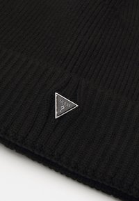 Gorro de punto acanalado negro con una etiqueta triangular plateada que presenta "GUESS" y un signo de interrogación en la parte frontal. Textura suave, diseño casual.