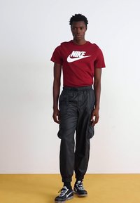 Camiseta roja de Nike con logotipo blanco, combinada con pantalones cargo de nailon negros y zapatillas negras con acentos blancos. El modelo está de pie sobre un suelo amarillo.