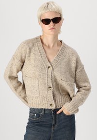 Bež pletena cardigan s v-izrezom, rebrastim robom, dvema sprednjima žepoma in velikimi gumbi. Nosi se čez temno modre kavbojke. Gladka tekstura.